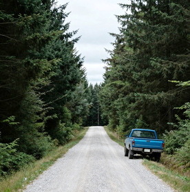 A blue pick-up truck parked along a gravel road through forest